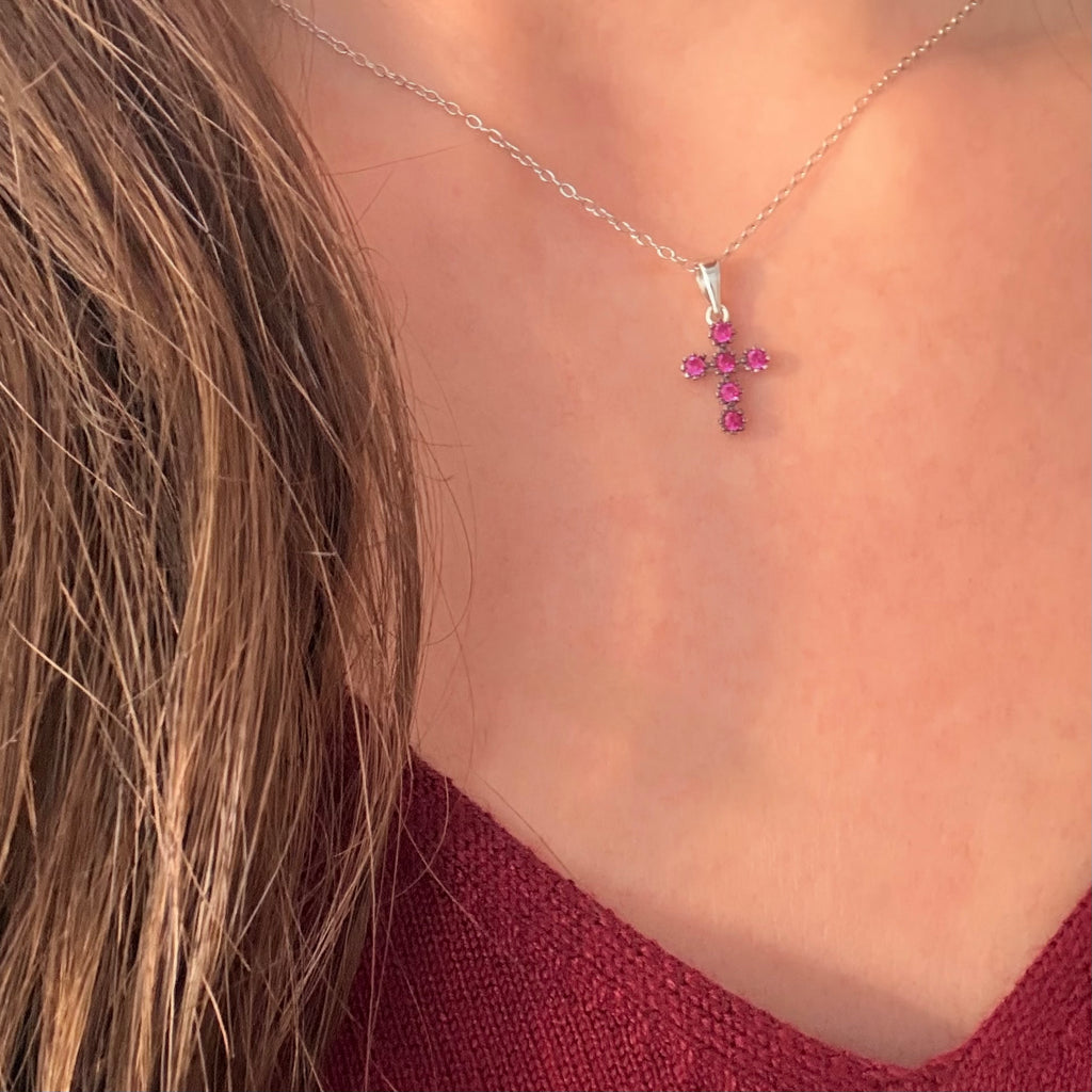 A close-up of a silver cross pendant necklace adorned with small pink gemstones, worn by a model with long light brown hair and a burgundy top.