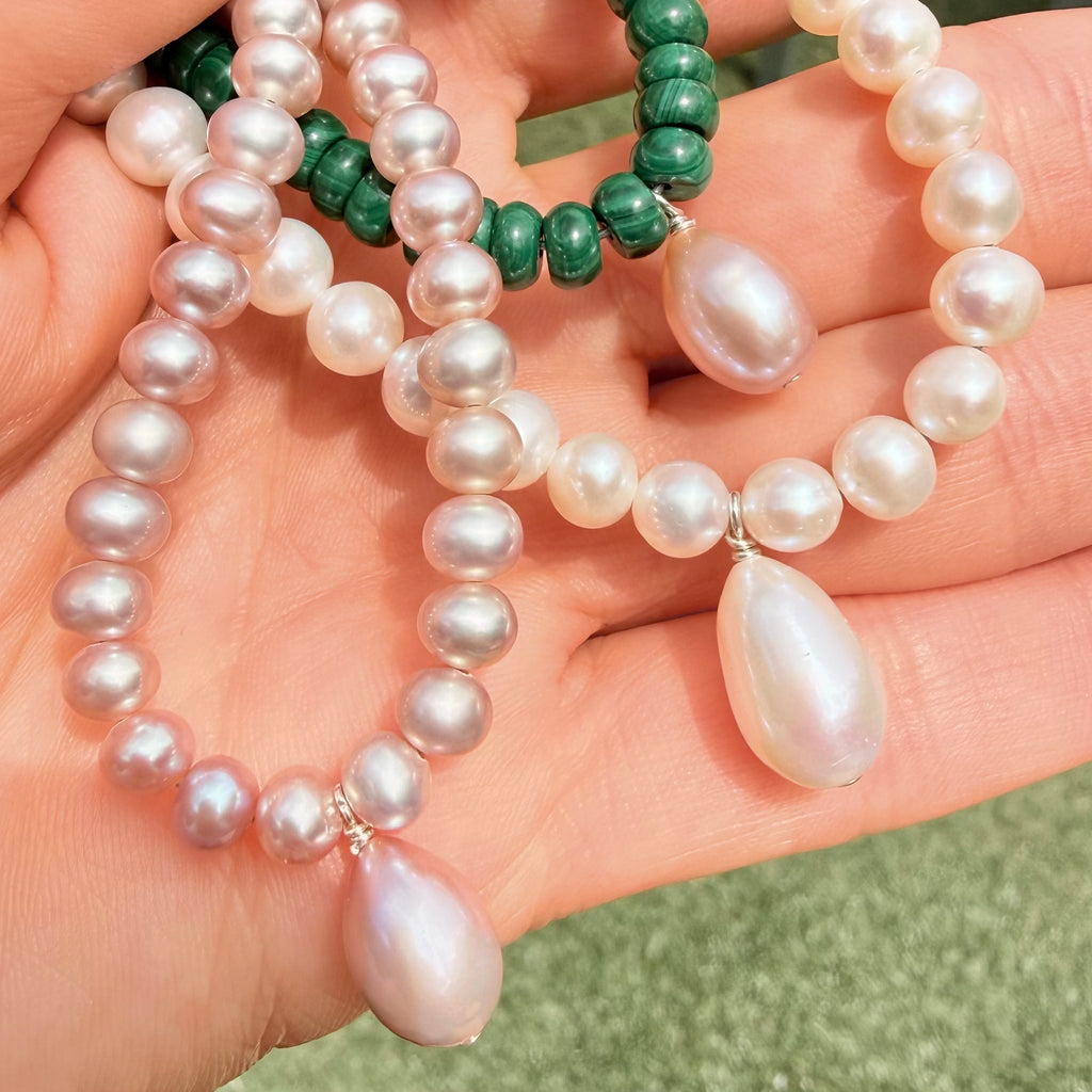 A close-up of a hand holding three pearl necklaces. One necklace features lustrous white freshwater pearls with sterling silver settings, one on lavender pink pearls, and one with malachite beads.