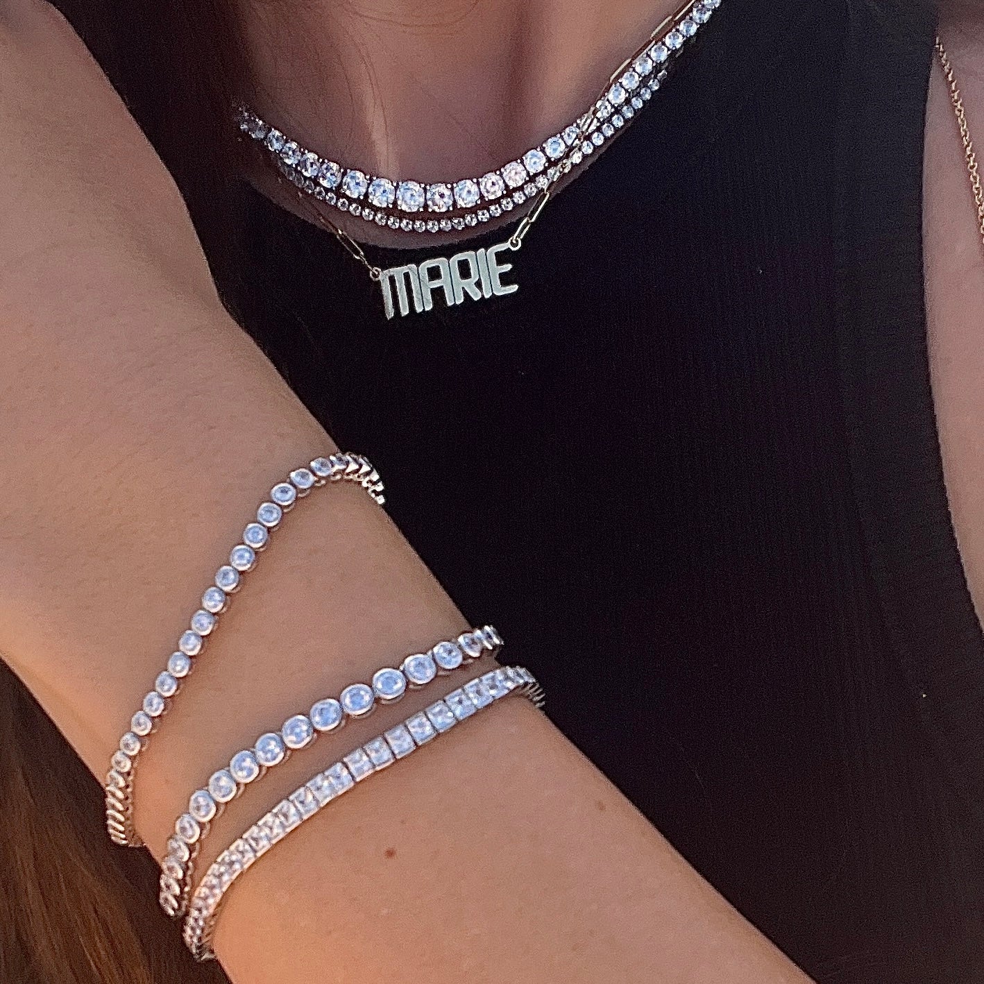 Close-up of a person's arm wearing multiple faux tennis diamond bracelets with a black top and tennis necklaces with a gold nameplate necklace 