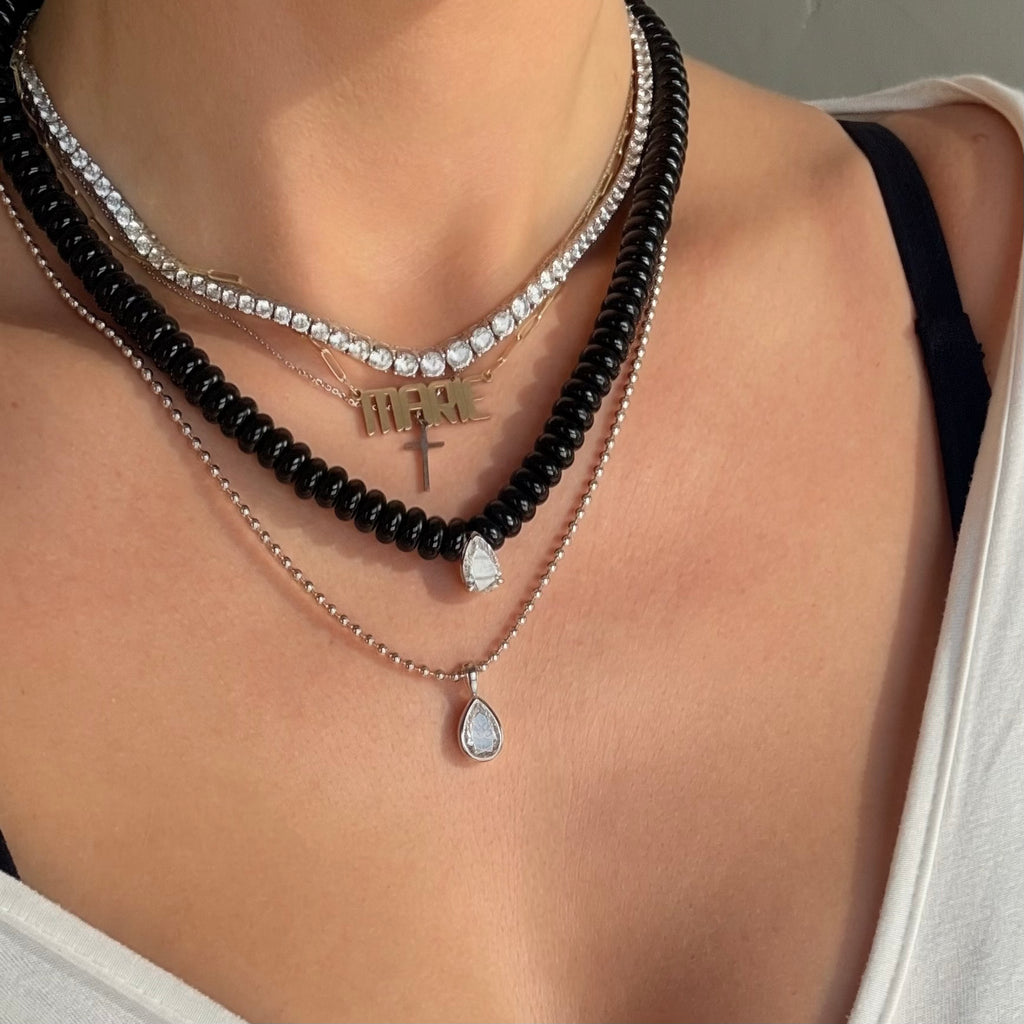A close-up of a person wearing a layered necklace set. The set includes a black agate beaded necklace, a graduated tennis necklace, a gold nameplate necklace, a silver mini cross pendant, and a delicate silver ball chain with a teardrop-shaped moissanite pendant.