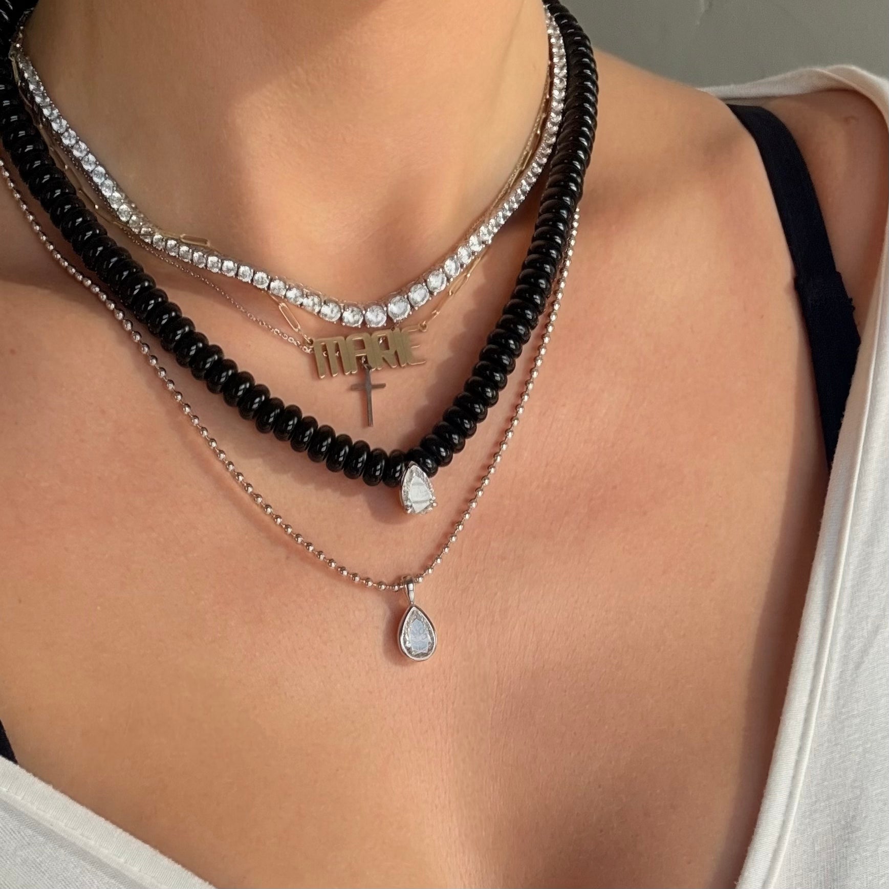 A close-up of a person wearing a layered necklace set. The set includes a black agate beaded necklace, a graduated tennis necklace, a gold nameplate necklace, a silver mini cross pendant, and a delicate silver ball chain with a teardrop-shaped moissanite pendant.