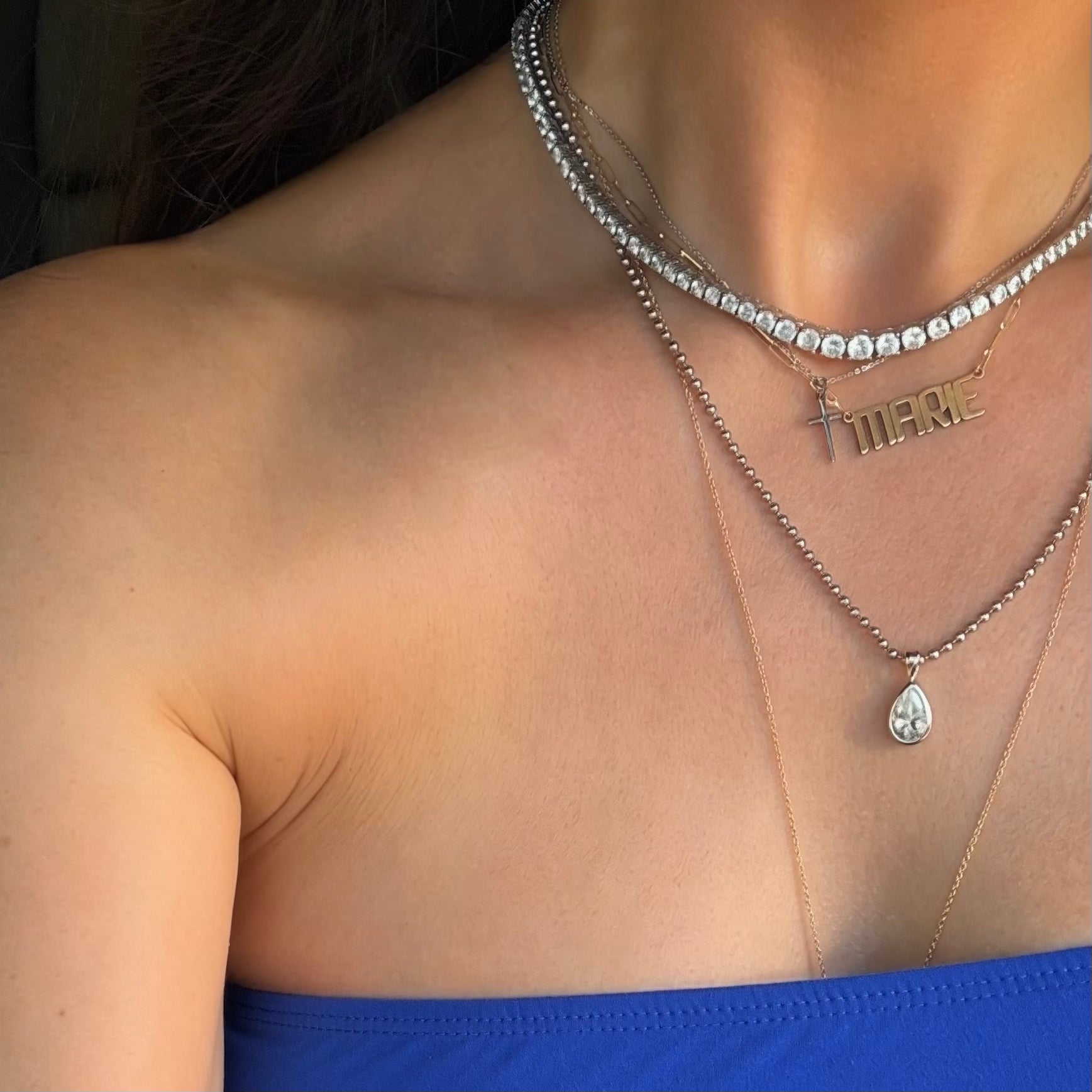 Layered silver and gold necklaces with a tennis necklace, a cross necklace, a nameplate, and a teardrop pendant necklace.
