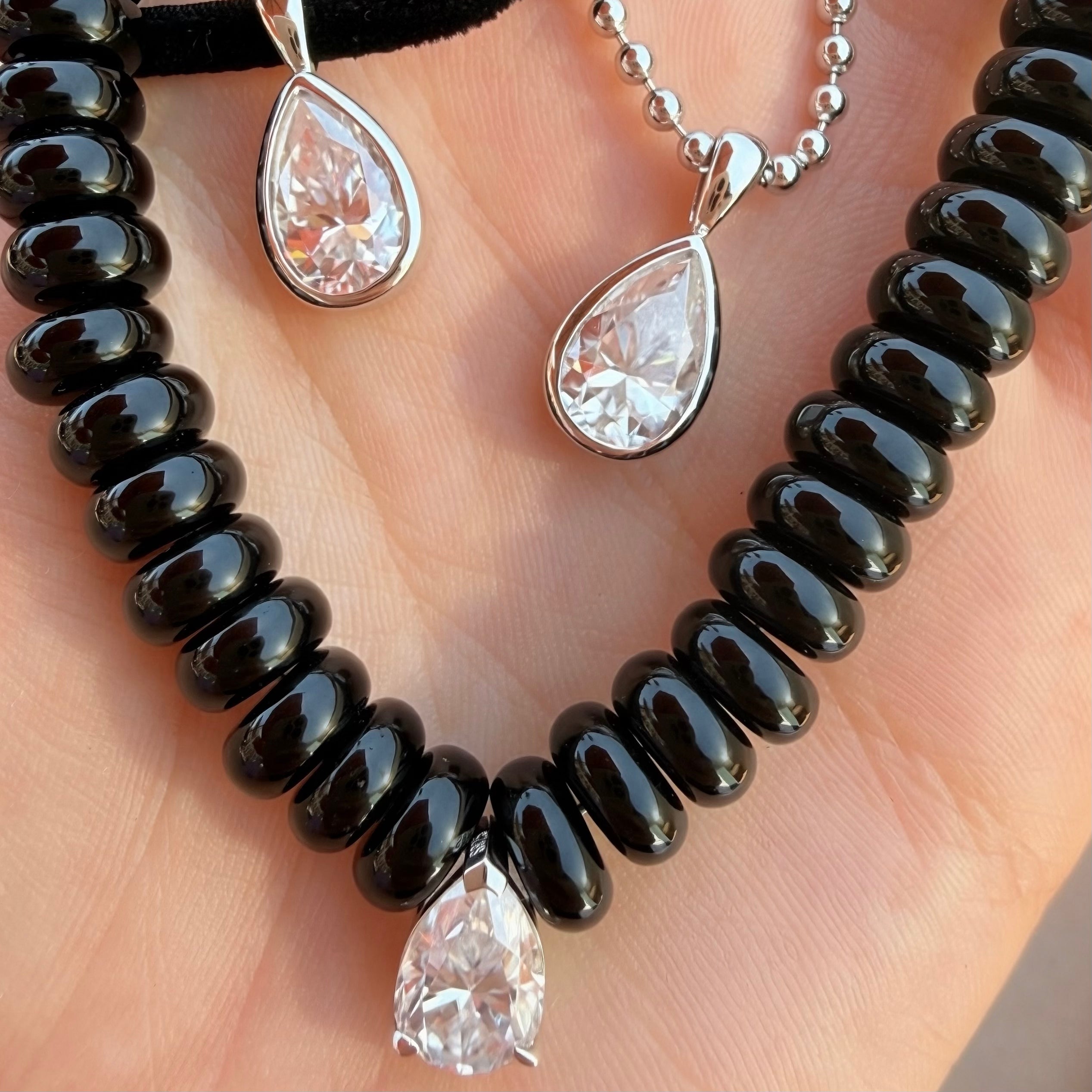 A close-up of jewelry displayed on a hand, featuring a black agate beaded necklace with a teardrop-shaped moissanite pendant, and a silver ball-chain necklace with a similar pendant.