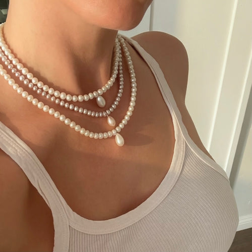 A close-up of a person wearing a layered pearl necklace with three strands of white pearls and three teardrop-shaped pearl pendants, styled with a white ribbed tank top.