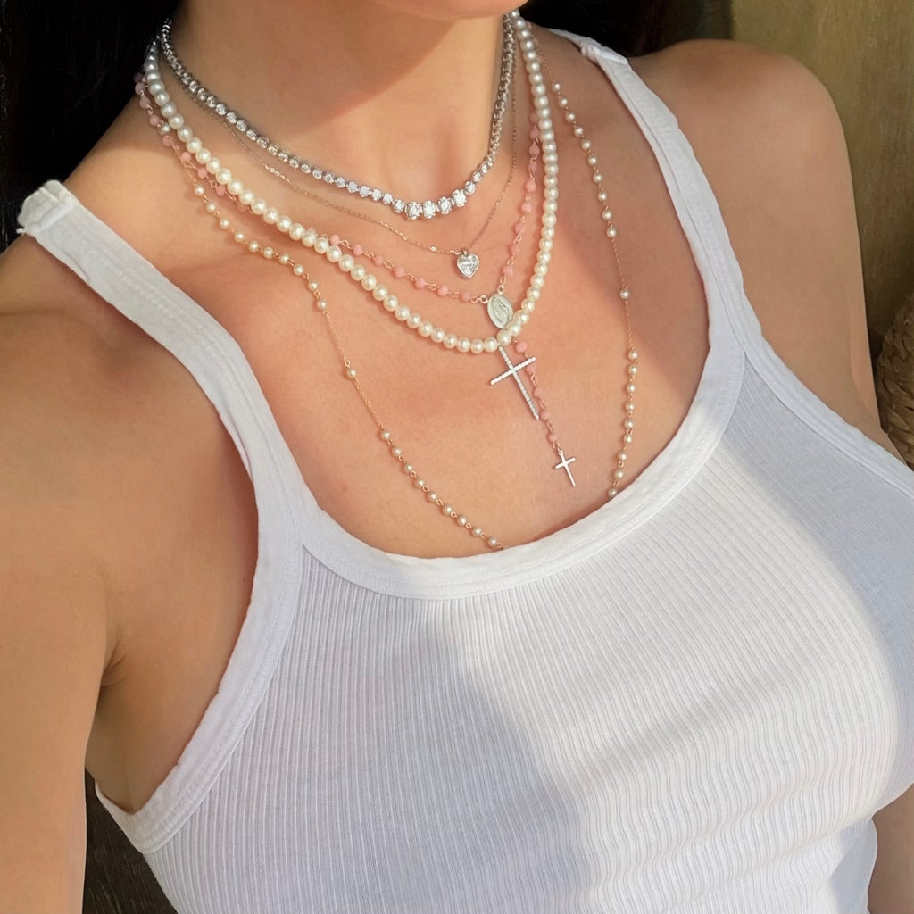 Pave cross pearl necklace layered with a tennis necklace, a heart pendant, and two pearl and pink rosary necklaces 