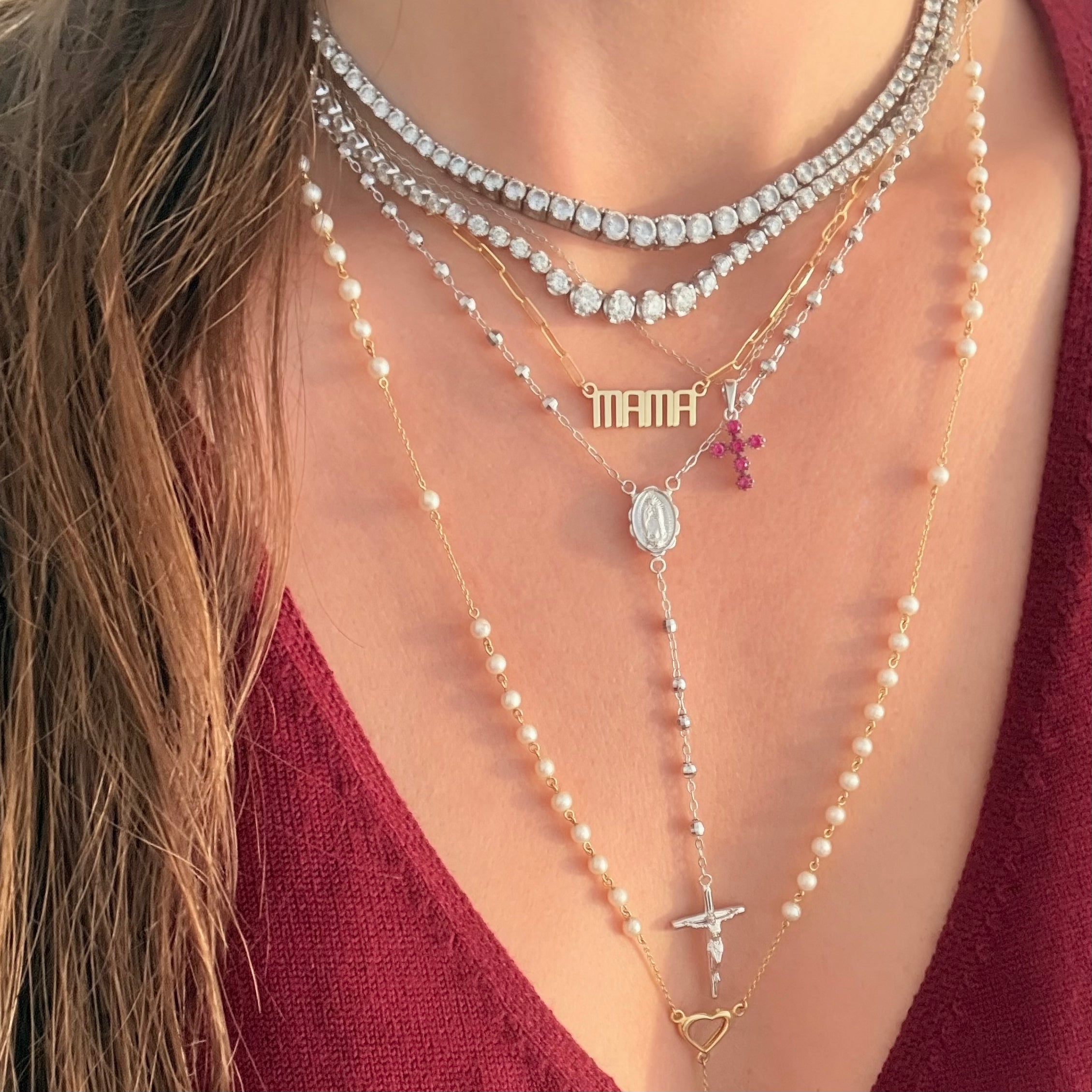 A close-up of a person wearing a layered necklace set. The set includes two tennis necklaces, a gold chain with a 'MAMA' nameplate and a small pink cross, a long pearl and gold rosary necklace with a heart-shaped pendant.