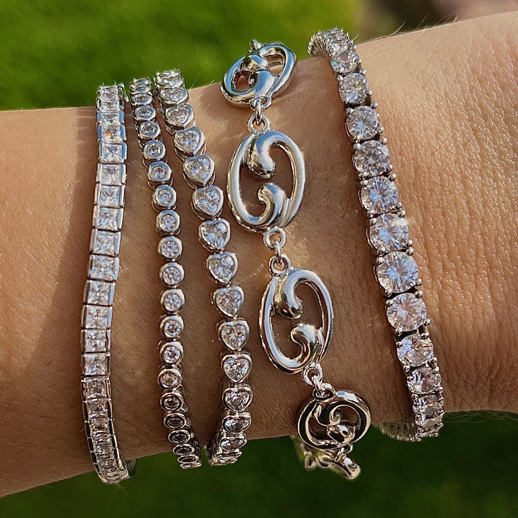 Stack of silver tennis bracelets on a wrist, with a heart bezel bracelet, a Fibonacci spiral bracelet, and a graduated tennis bracelet 