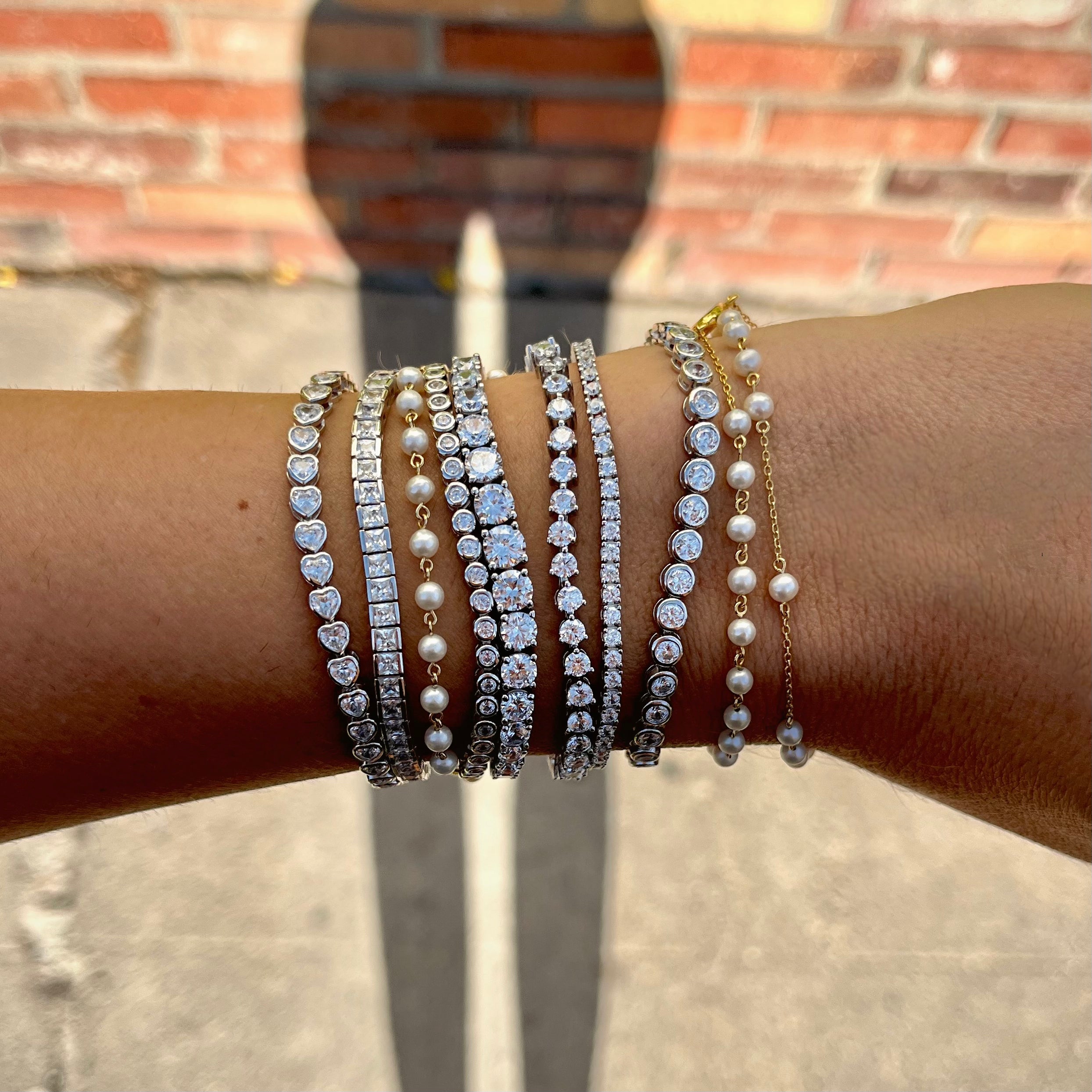 Multiple tennis bracelets and pearl bracelets on a wrist with a blurred brick wall background